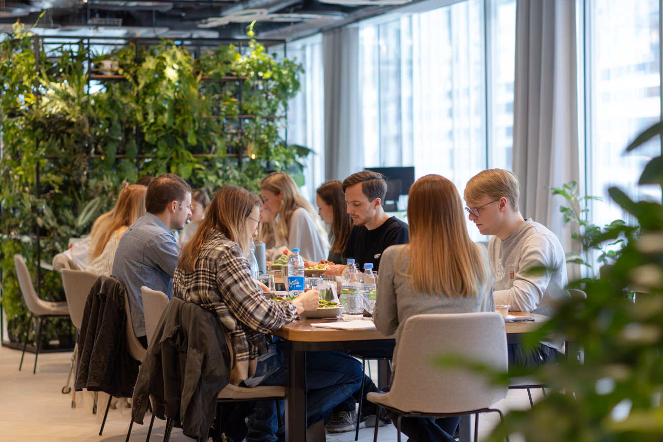 Mehrere junge Erwachsene sitzen gemeinsam an einem Tisch in einem modernen, grünen Raum und essen miteinander