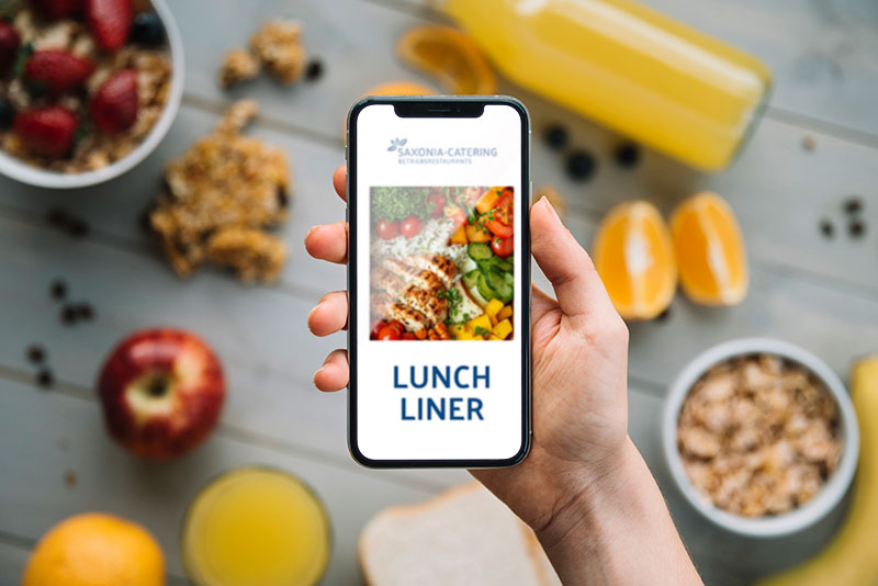 Eine Hand hält ein Smartphone mit der Lunch-Liner-App vor einem Frühstückstisch.